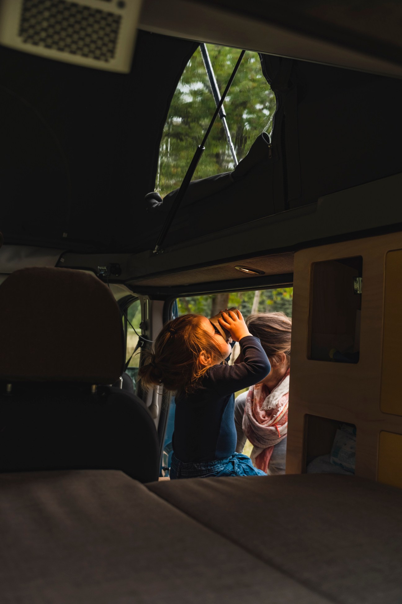Person looking through binoculars inside a camper van with roof window open showing green trees