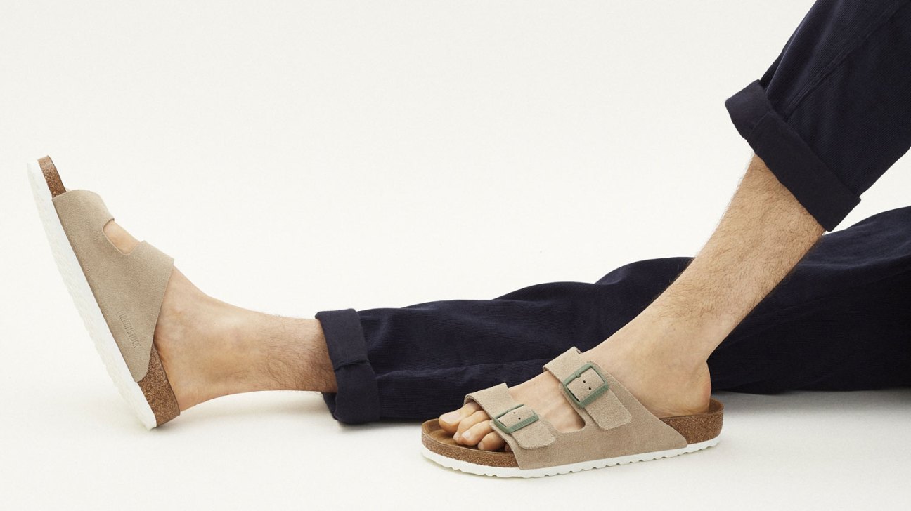 Person wearing beige Birkenstock Arizona sandals with two adjustable straps, sitting in dark clothing