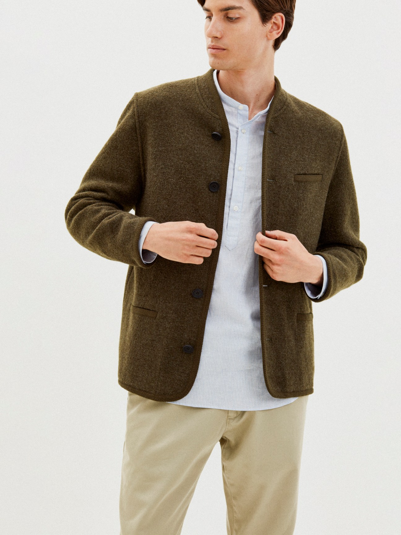 Man wearing olive green wool jacket with band collar over light blue striped shirt and beige trousers