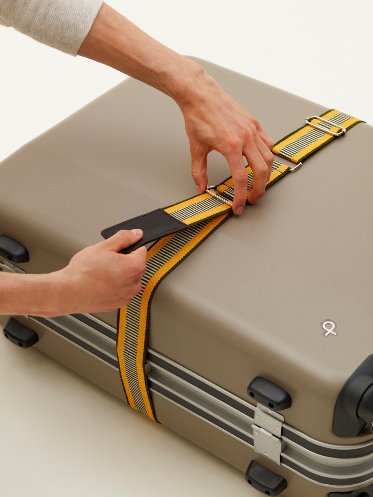 Hands fastening a yellow and gray striped luggage belt around a beige suitcase.
