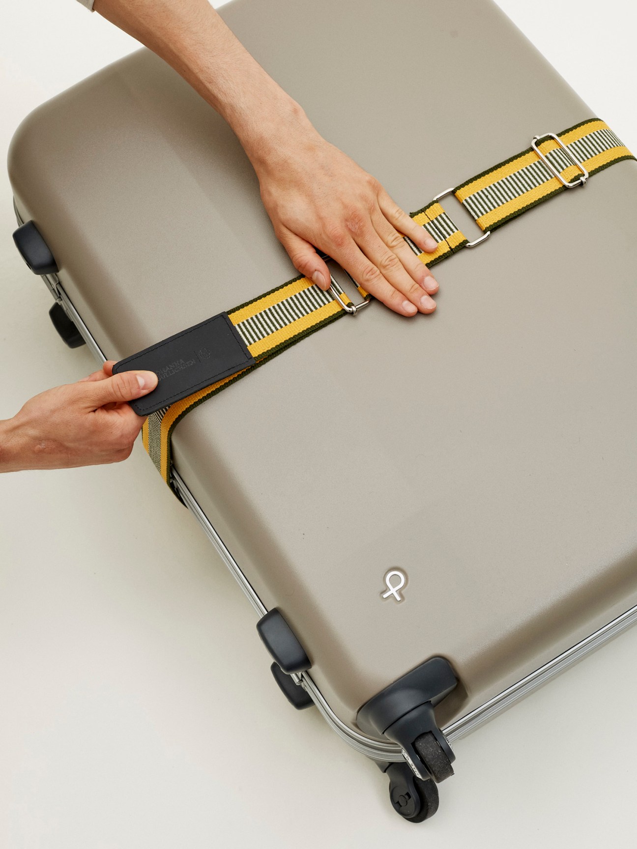 Hands securing a yellow and black luggage belt around a beige suitcase.