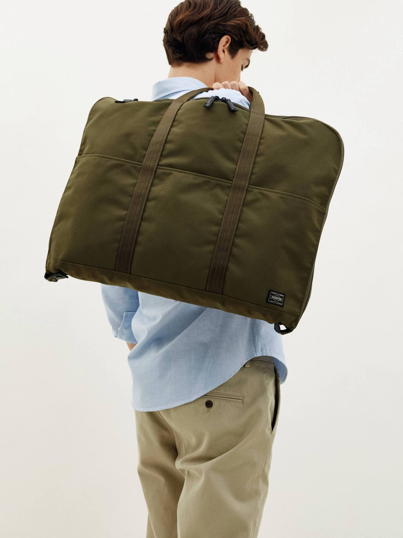 Man carrying olive Porter x Monocle suit bag with dual straps over shoulder wearing light blue shirt