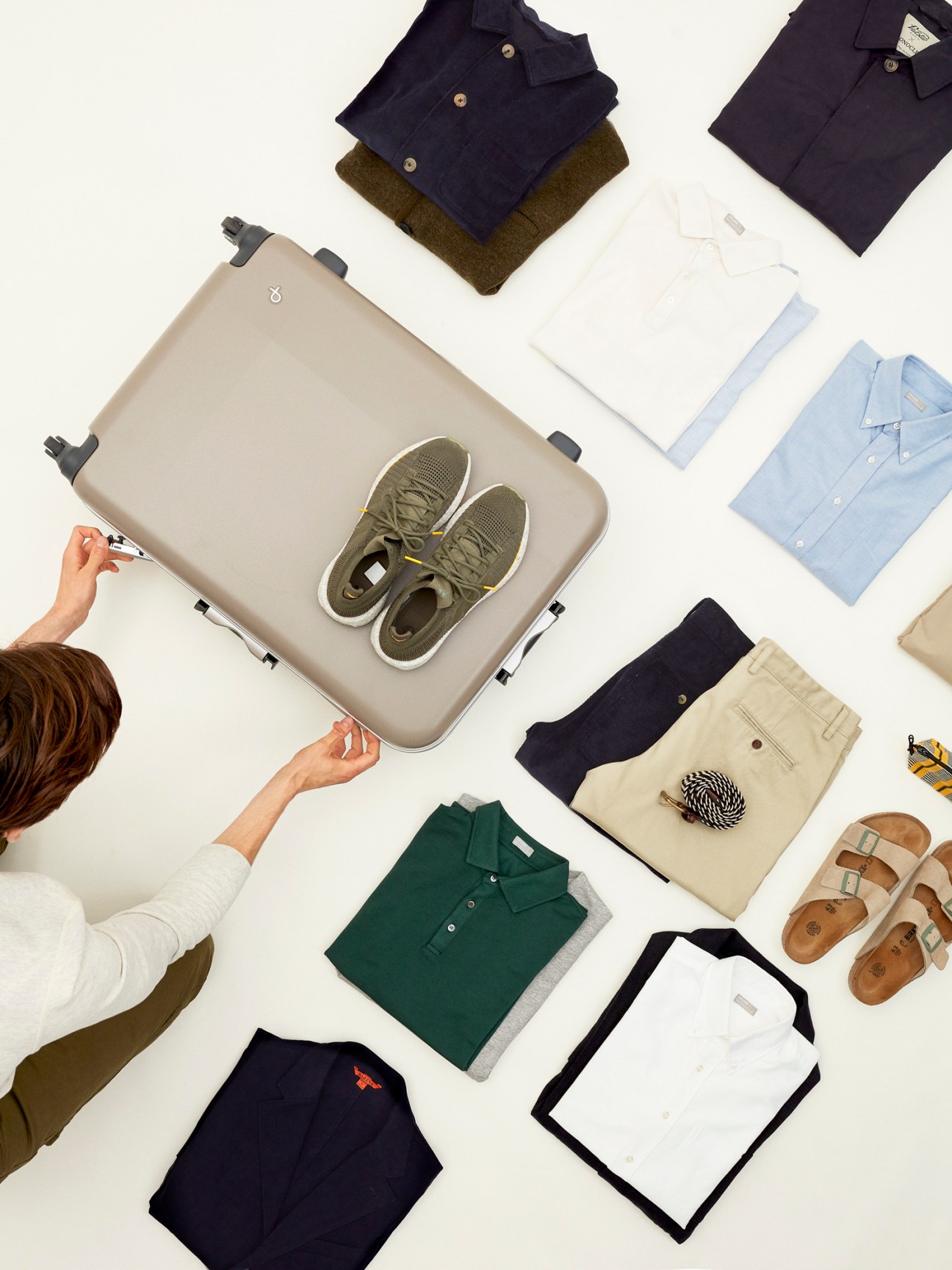 Person packing olive green sneakers into beige suitcase surrounded by neatly arranged clothing items