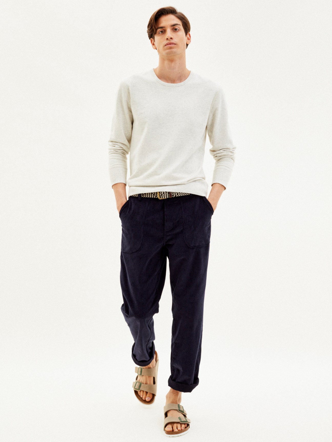 Man wearing oatmeal round-neck sweatshirt with navy trousers, patterned belt and sandals on white background