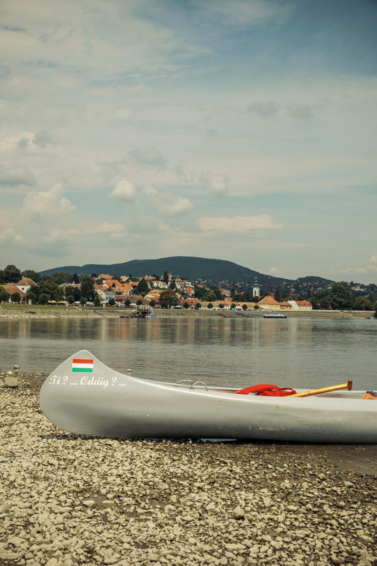 White canoe with Hungarian flag and text on a pebble beach beside the Danube River with town and mountains beyond.
