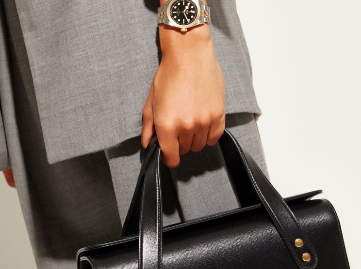 Person wearing gray blazer holding black Gucci bag with gold horsebit detail and Tudor Black Bay watch.