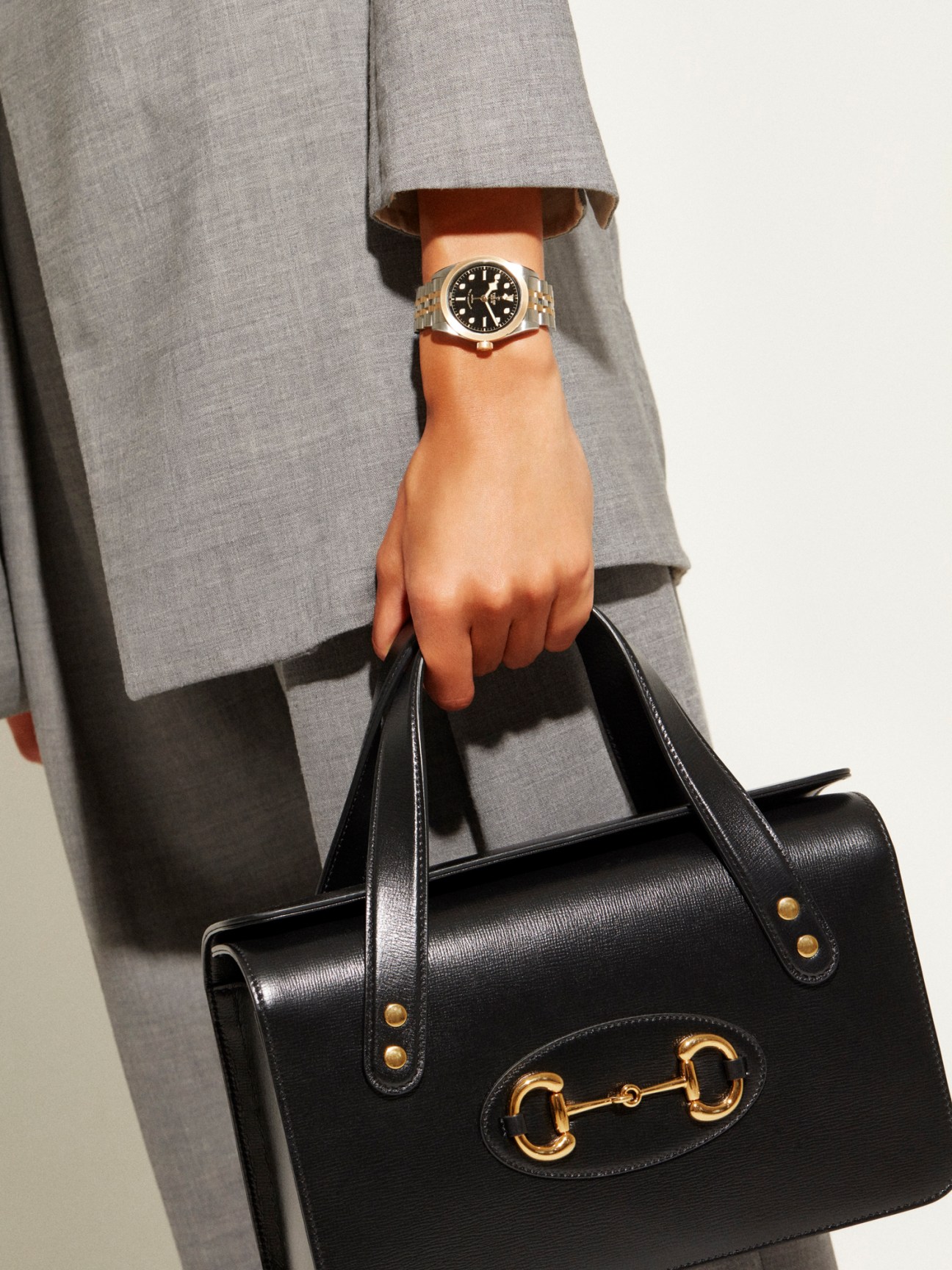 Person wearing gray blazer holding black Gucci bag with gold horsebit detail and Tudor Black Bay watch.