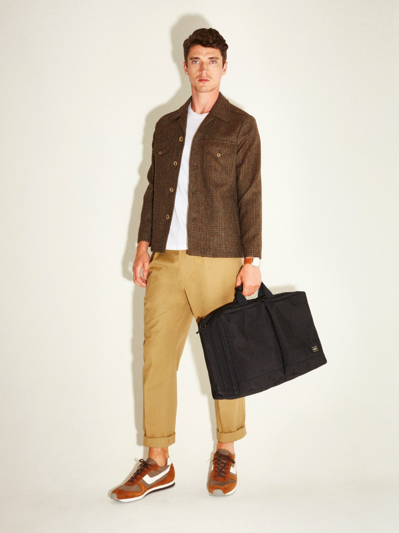 Man wearing brown Finamore jacket, khaki Drake's trousers, brown Reproduction of Found trainers, and holding a black Porter bag.