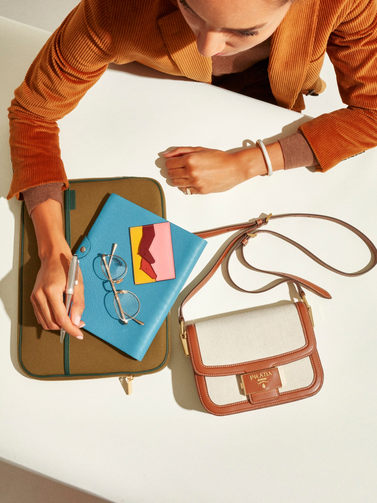 Person wearing Officine Générale jacket and Lardini rollneck jumper displaying Prada bag, Hermès notebook and card case, and Montblanc pen on white surface.