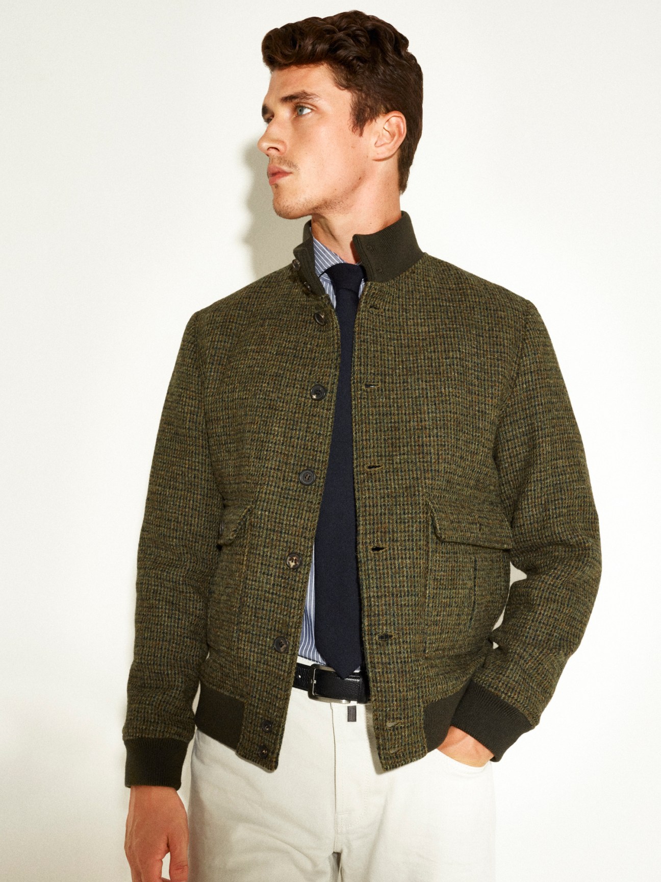 Man wearing olive green Valstar jacket with striped shirt, navy tie and white trousers against white background.