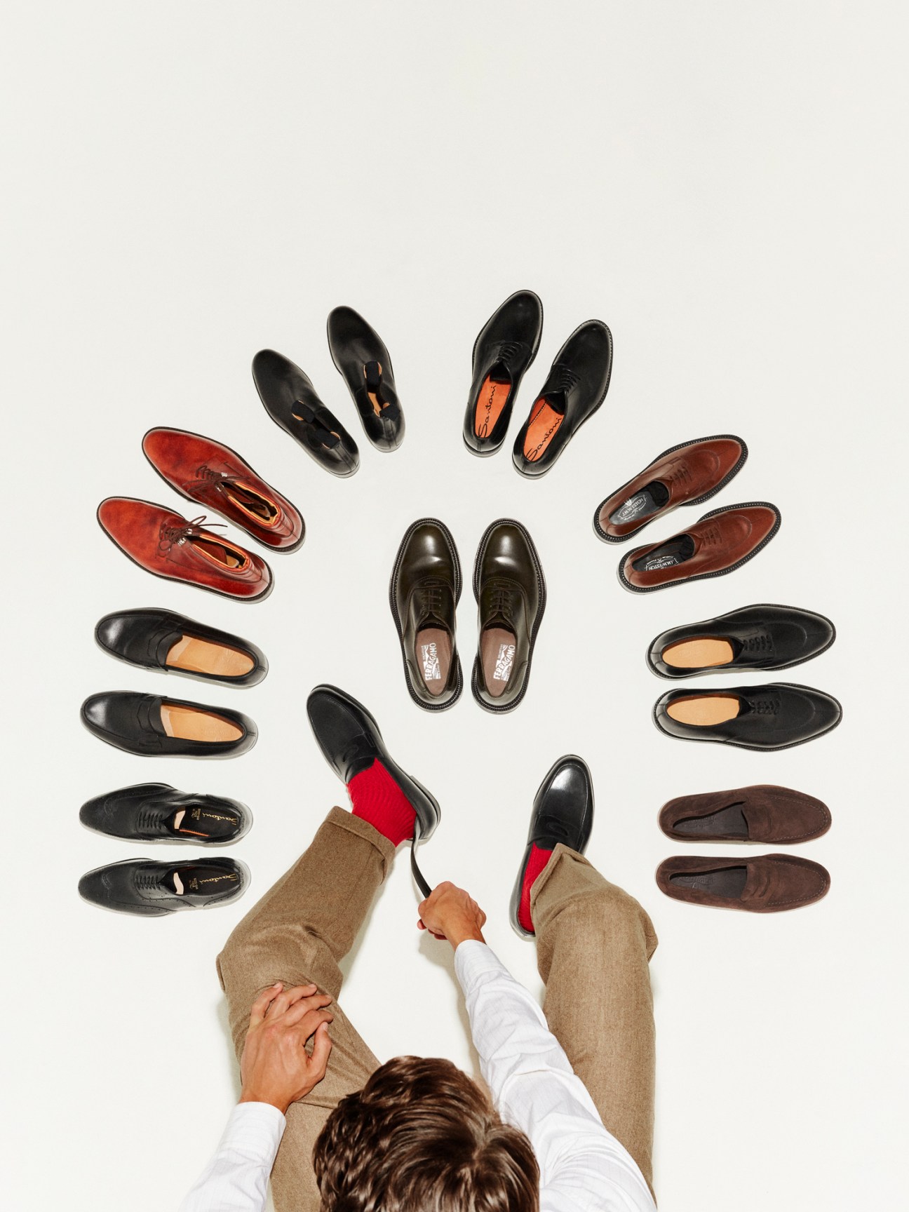 Overhead view of a person wearing tan trousers with red socks and black shoes, surrounded by nine pairs of dress shoes from luxury brands.