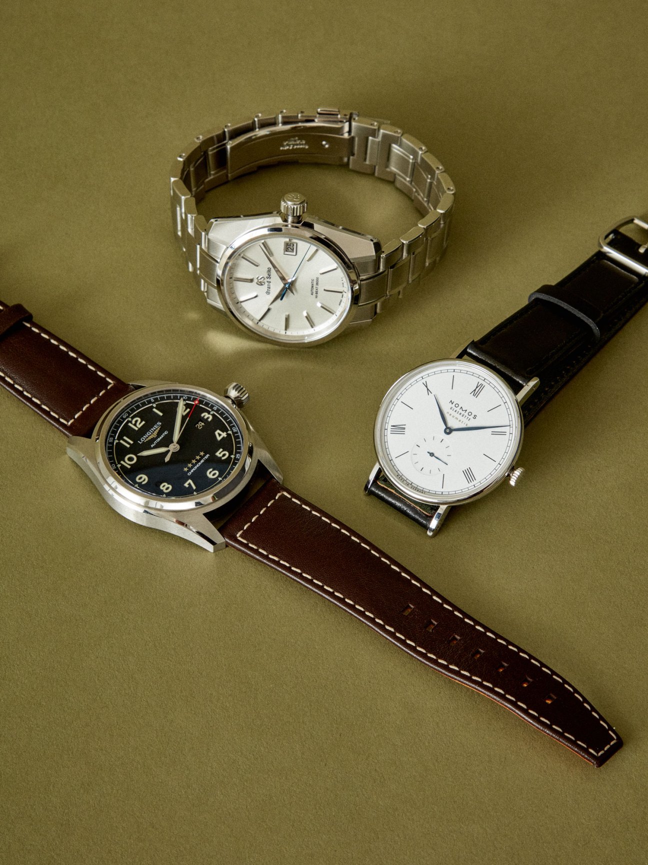Three luxury watches displayed on olive green background - one with steel bracelet, two with leather straps