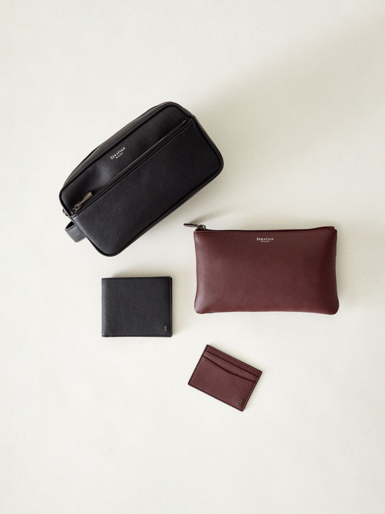 Black leather toiletry bag, burgundy pouch, black wallet and burgundy card holder by Serapian arranged on beige surface