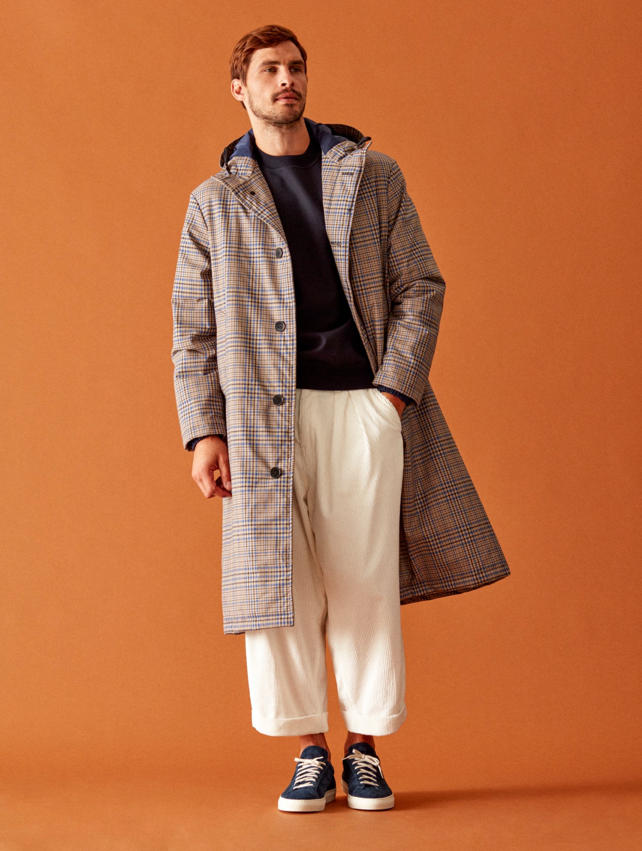 Man in plaid overcoat, black sweater, and cream trousers poses against orange background wearing navy sneakers