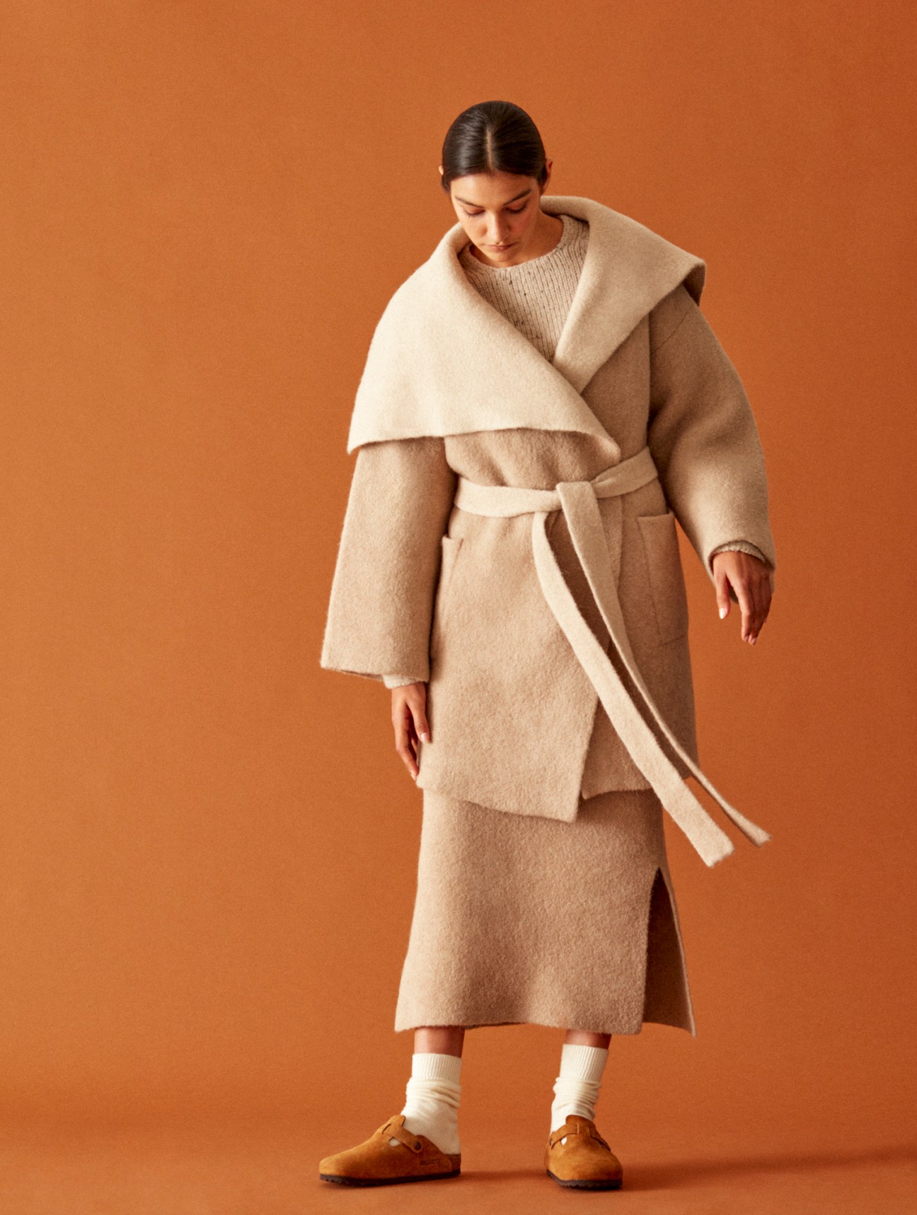 Woman in oversized beige wool coat with dramatic collar and belt, paired with knit sweater and midi skirt