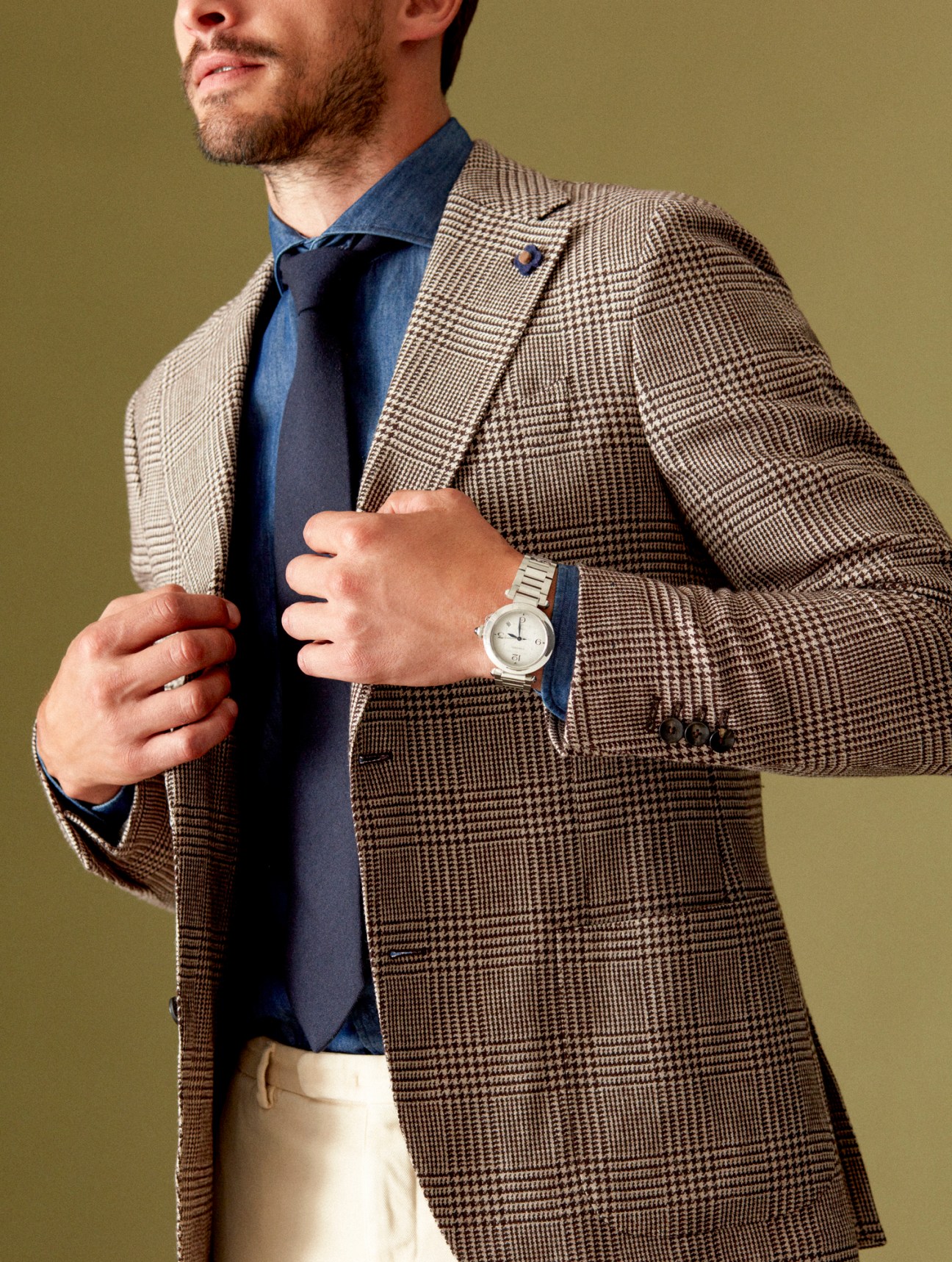 Man in brown houndstooth blazer with navy shirt and tie, wearing watch, adjusting jacket lapel