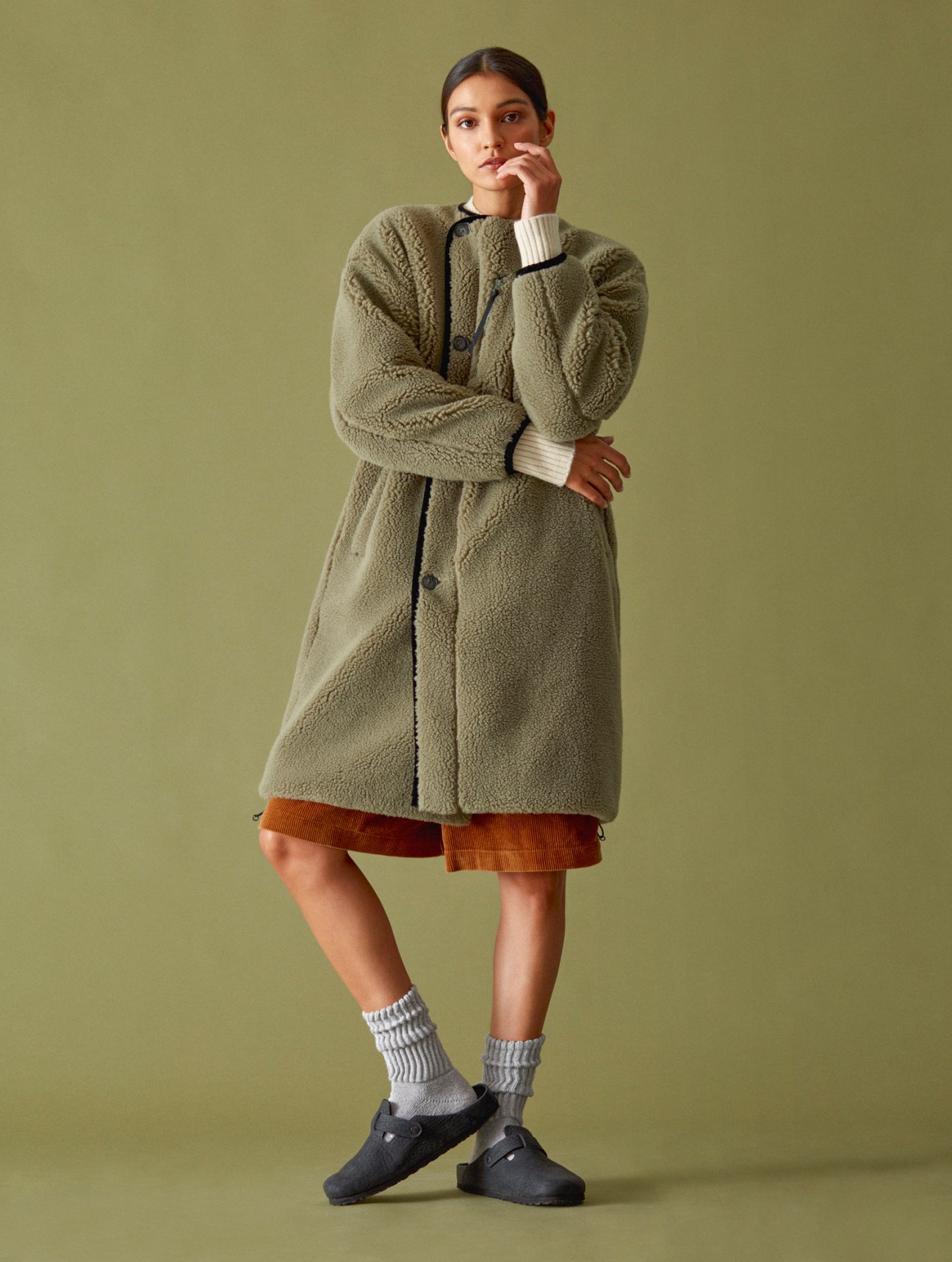 Woman in olive green fleece coat over rust corduroy skirt with striped socks and clogs against neutral background