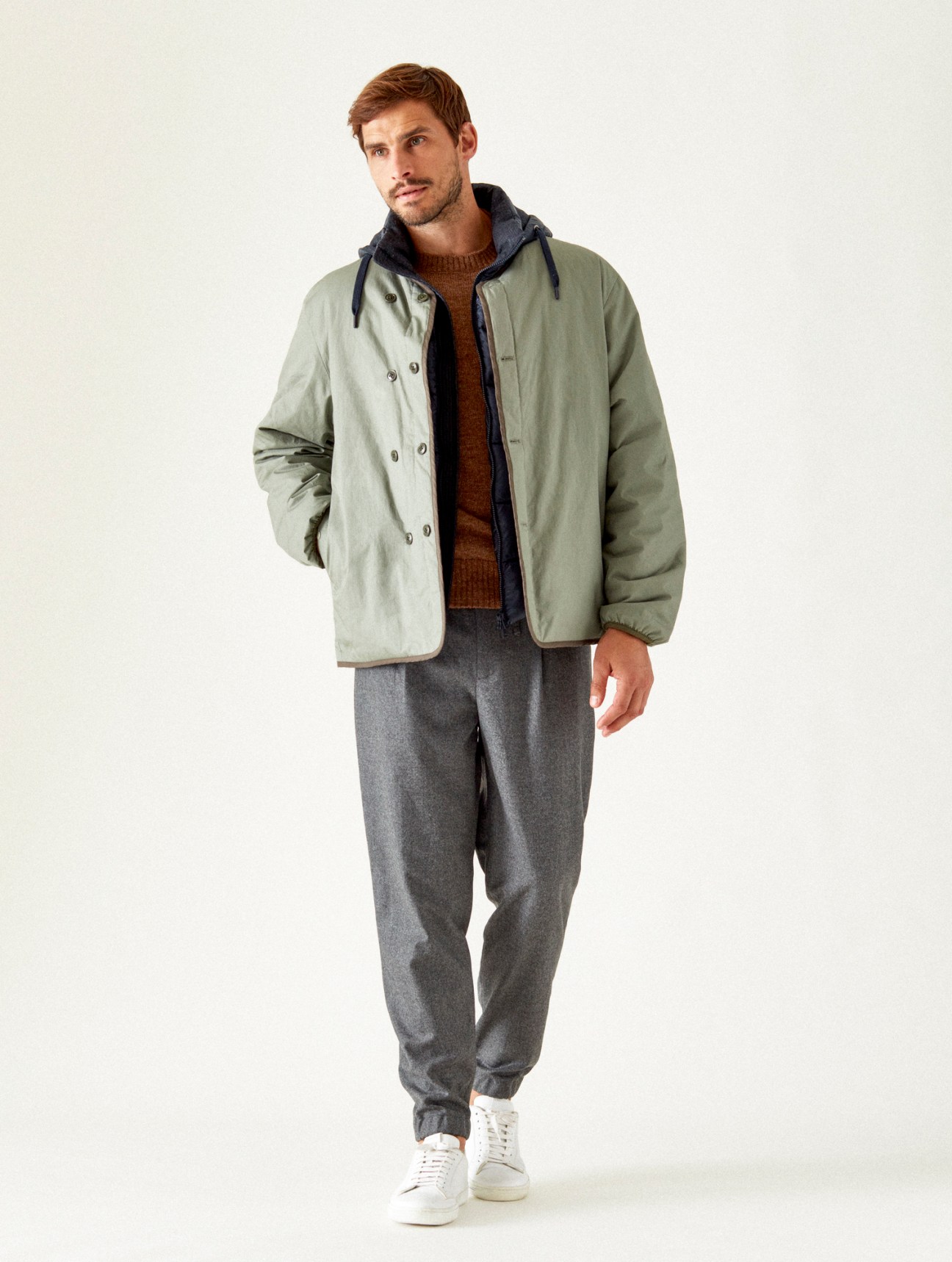 Man in sage green jacket layered over brown sweater and grey trousers, looking away from camera