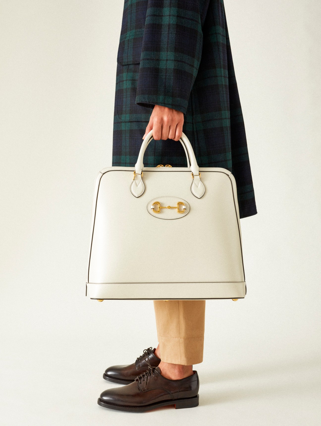 Person wearing navy plaid coat and beige trousers, holding cream leather handbag with gold hardware, brown dress shoes