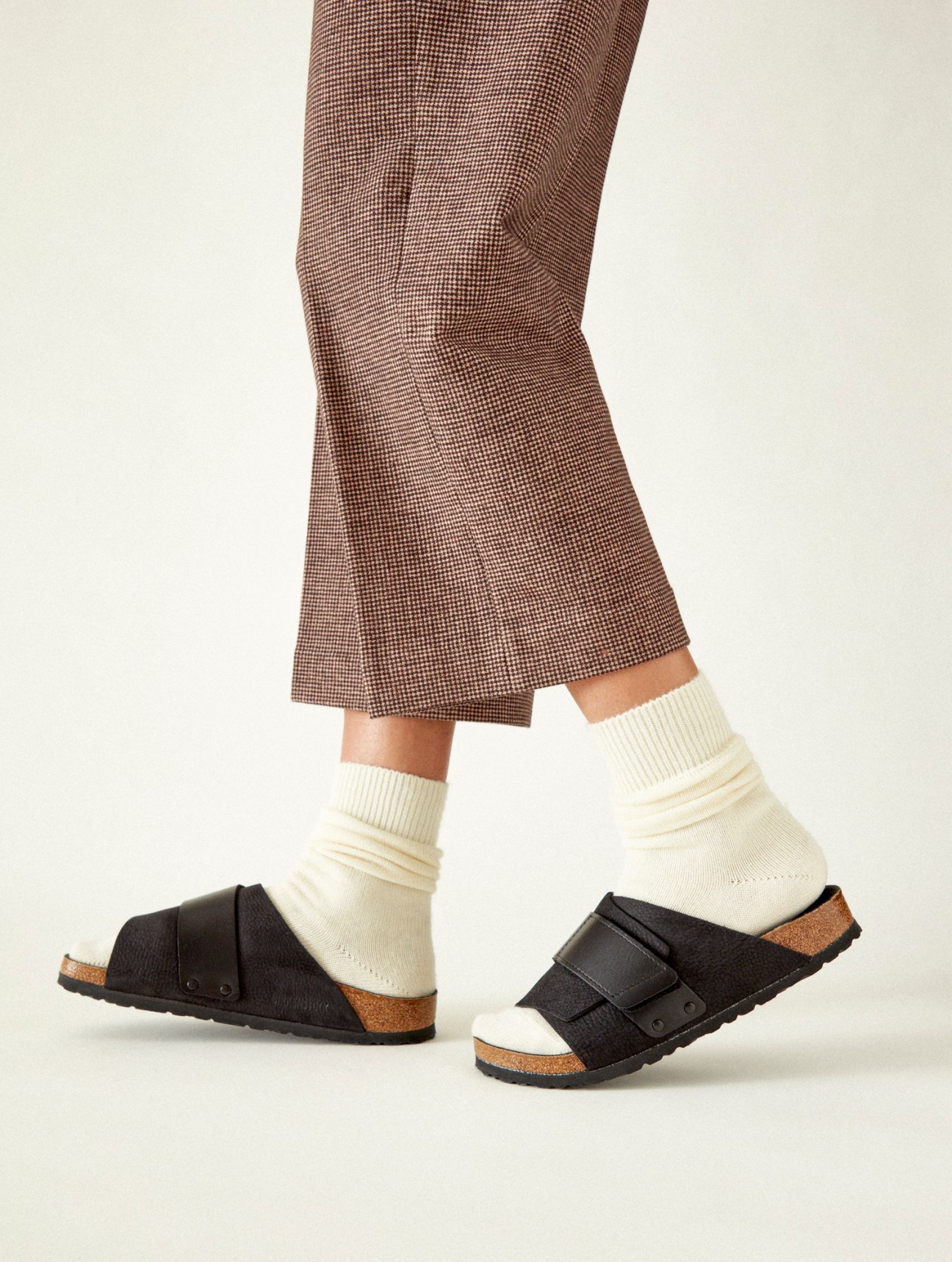 Person wearing brown houndstooth trousers with black Birkenstock sandals and cream ribbed socks