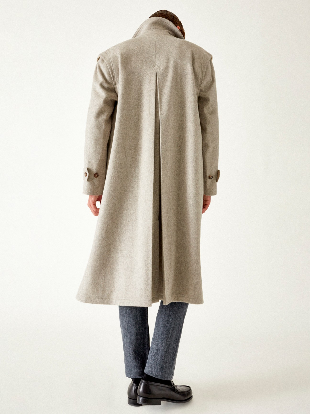 Model wearing long beige coat over dark jeans with leather dress shoes, shown from behind