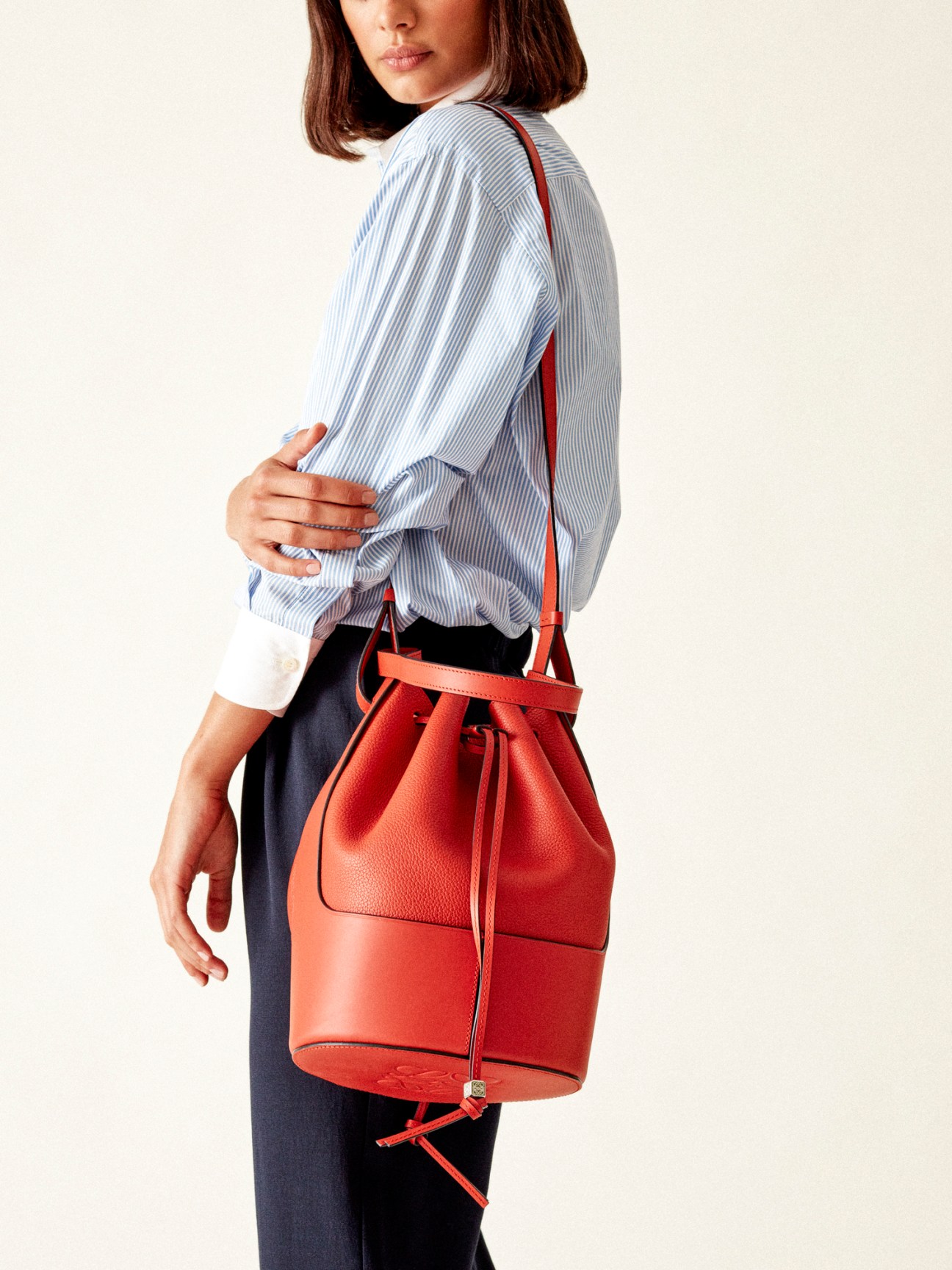Woman wearing blue striped shirt and dark trousers carrying red leather bucket bag