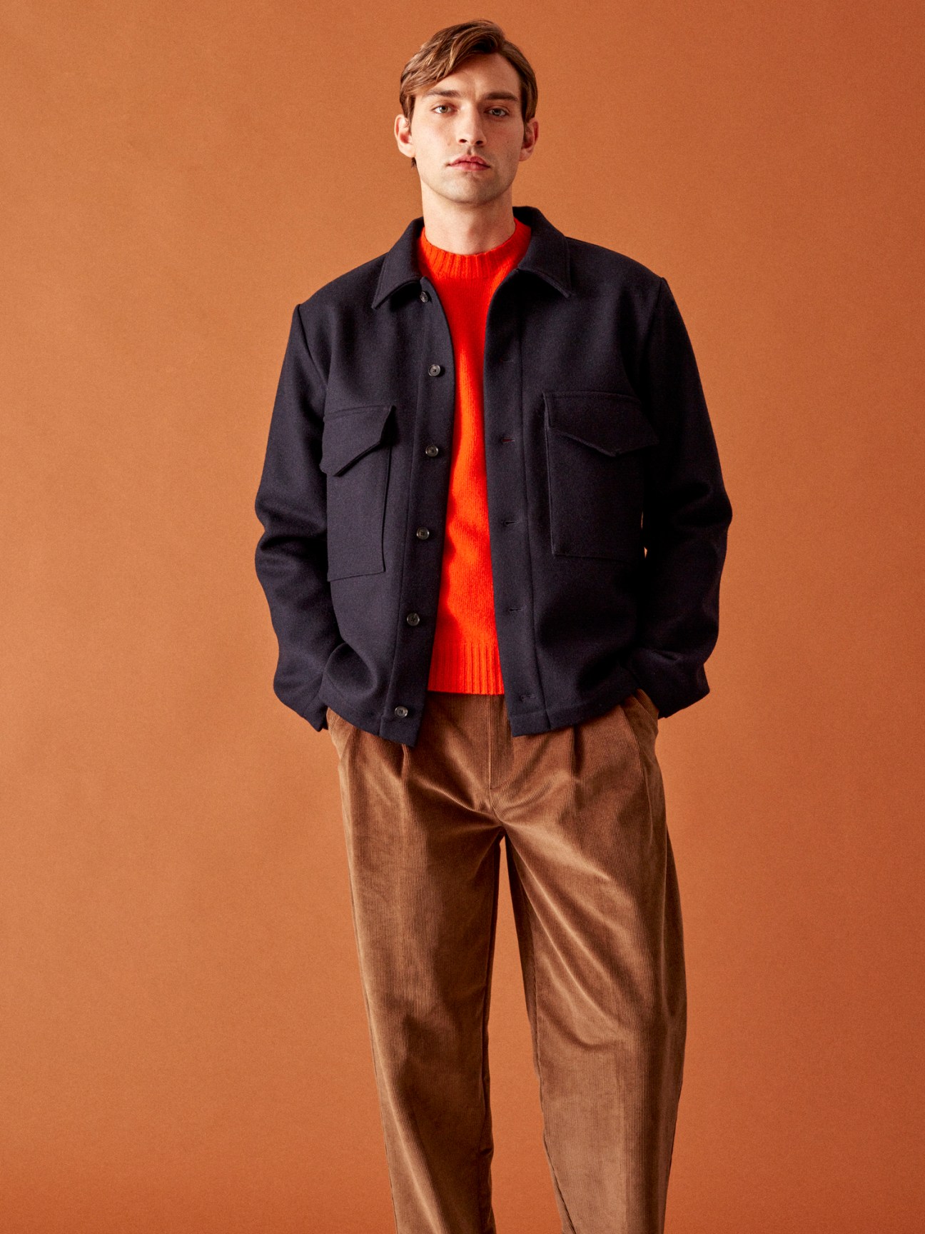 Model wearing navy jacket over red sweater and brown corduroy trousers