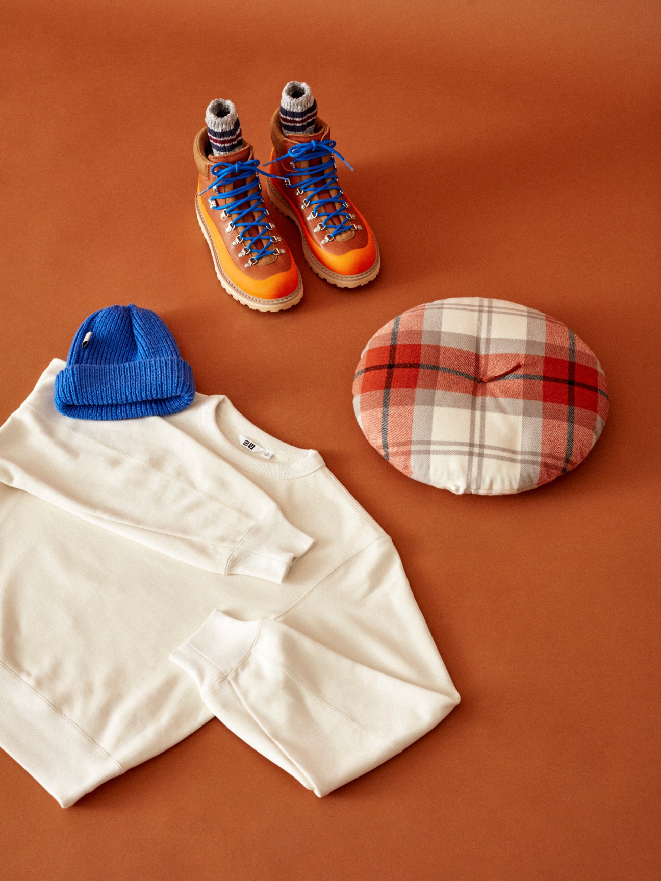 Orange hiking boots with striped socks, cream sweatshirt, blue beanie, and plaid beret arranged on rust background