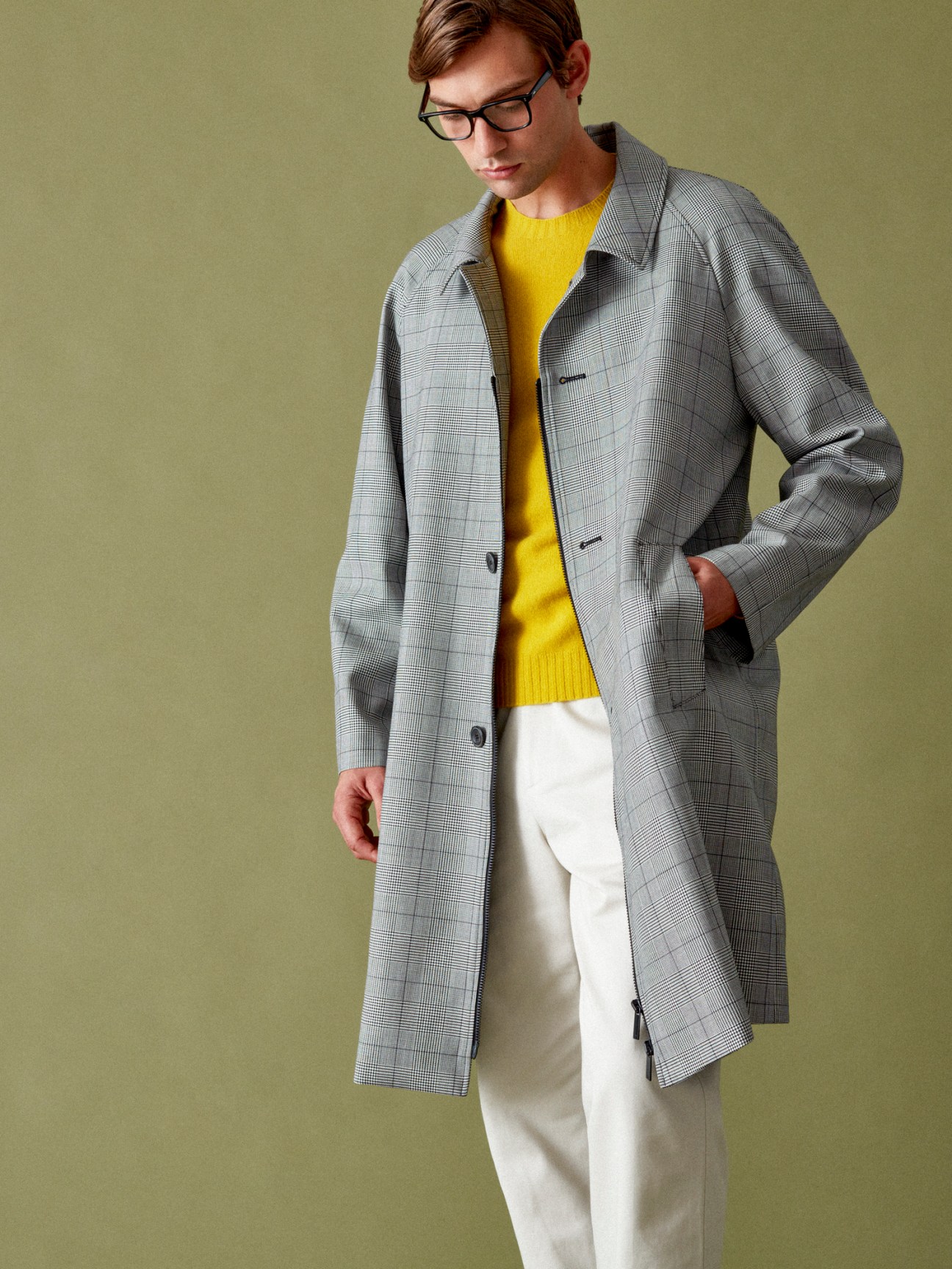 Man in glasses wearing grey plaid overcoat over yellow sweater and white trousers against olive background