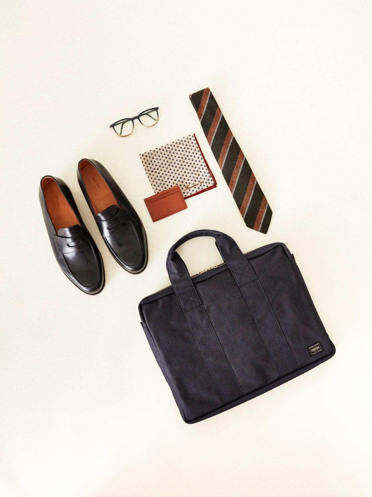 Flat lay of men's accessories including black leather loafers, briefcase, striped tie, glasses and pocket square