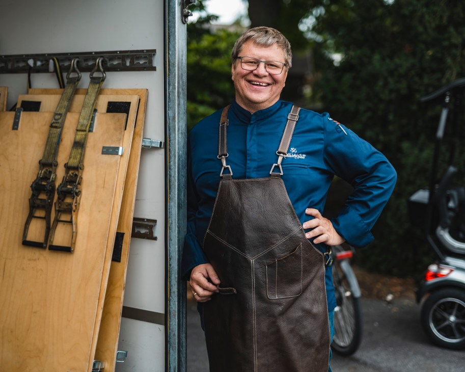 Chef and grill expert Sven Schurig wearing apron and glasses, smiling while standing next to outdoor equipment