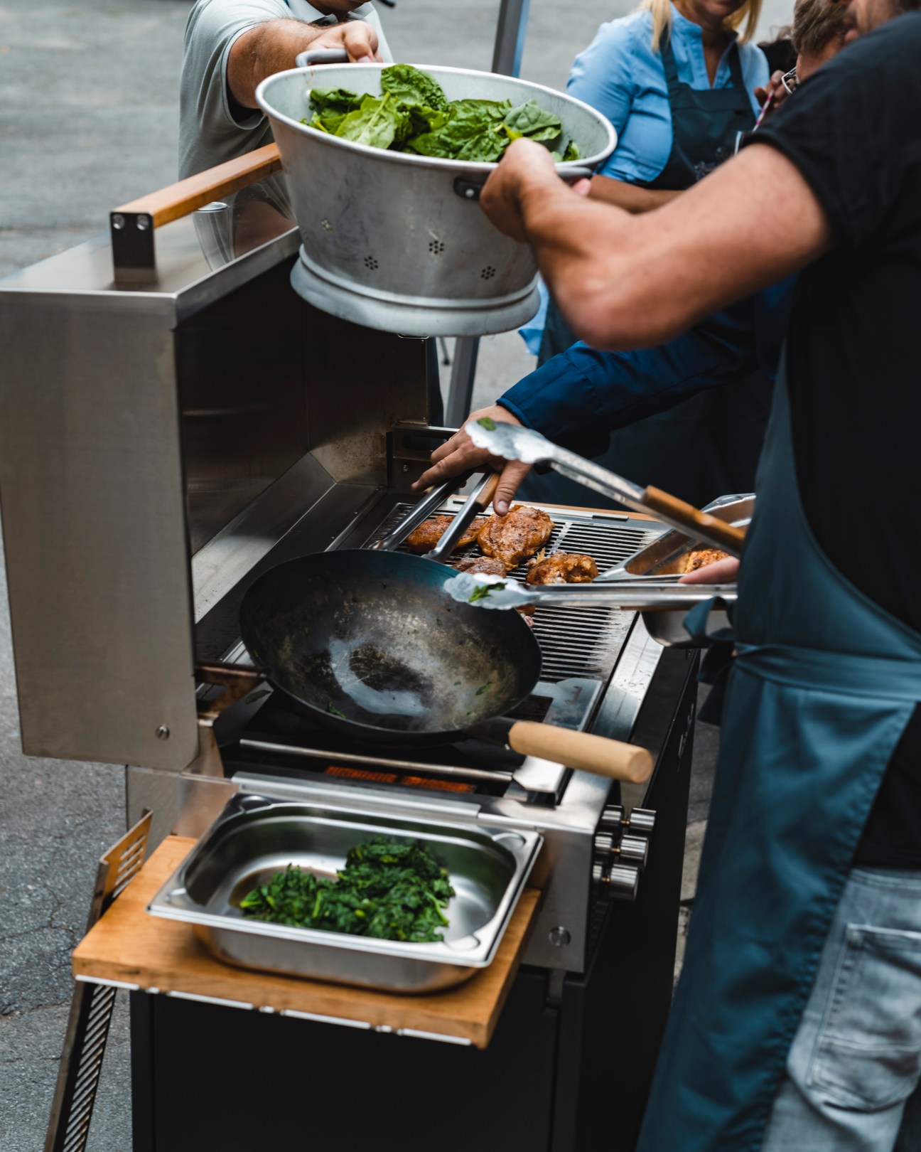 Person operating a Flammkraft grill, handling leafy greens in a colander over the cooking surface