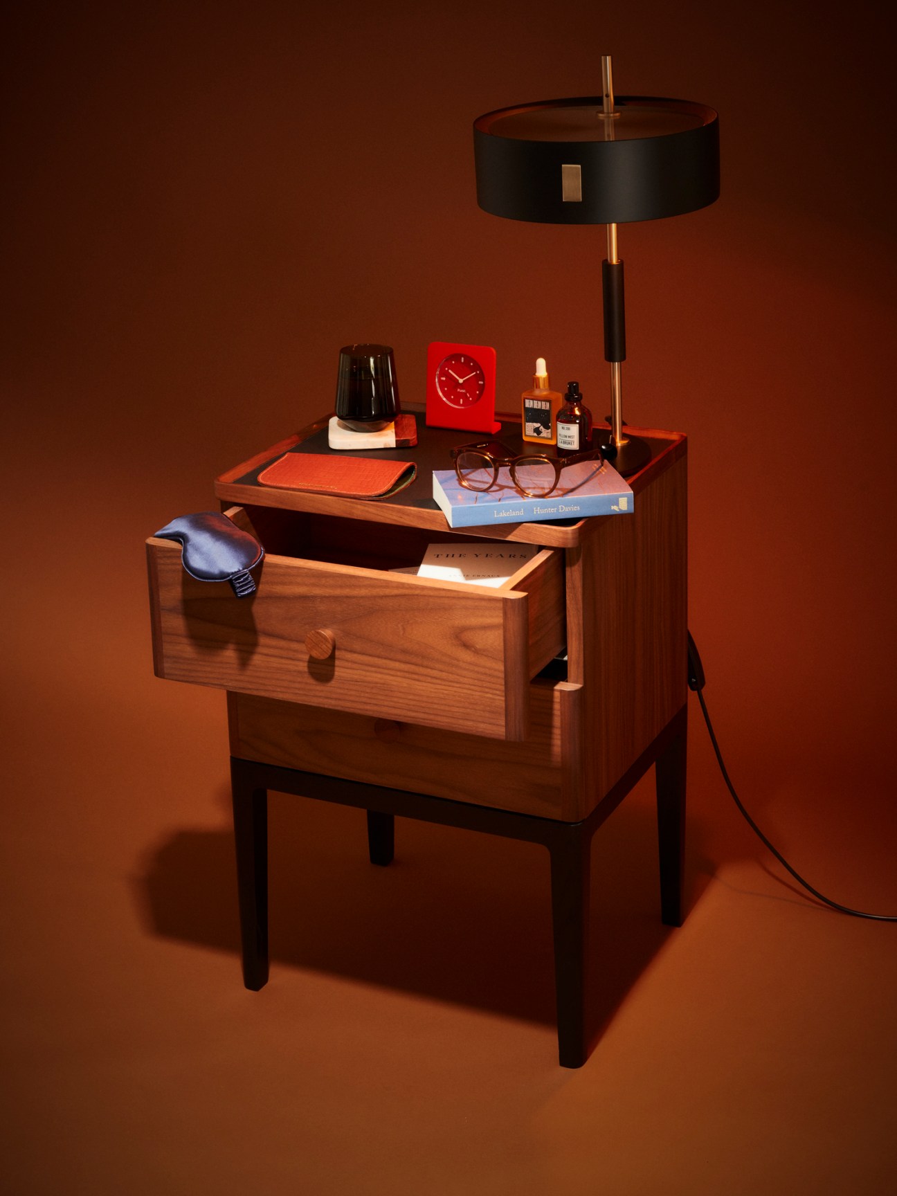Mid-century walnut bedside table with open drawer, lamp, clock, glasses, books and small bottles on top