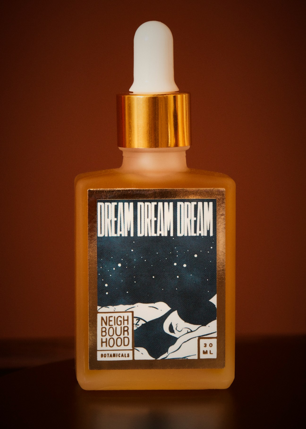 Glass dropper bottle of face oil with gold cap and illustrated label showing starry night sky