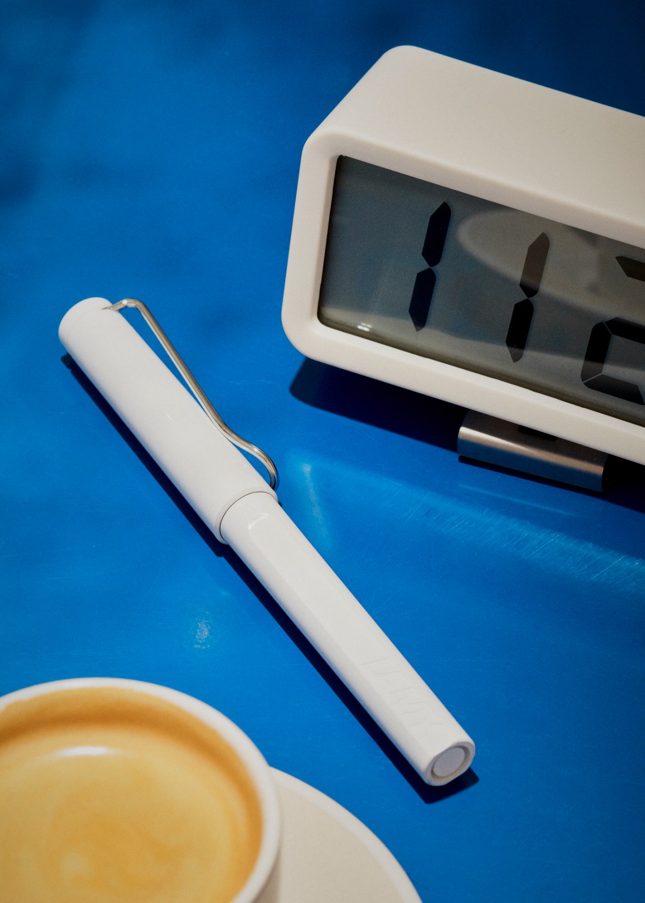 White Lamy pen with clip on blue surface next to digital clock and coffee cup