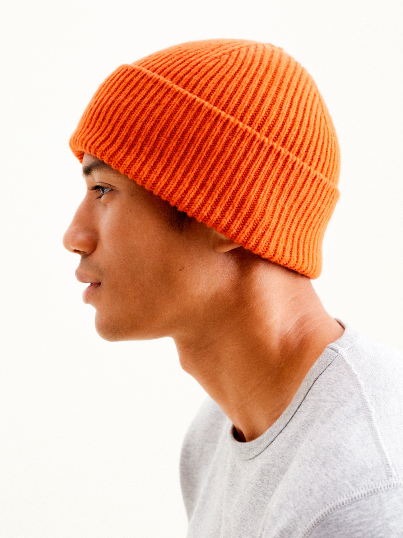 Person in profile wearing bright orange ribbed knit beanie and light grey shirt against white background