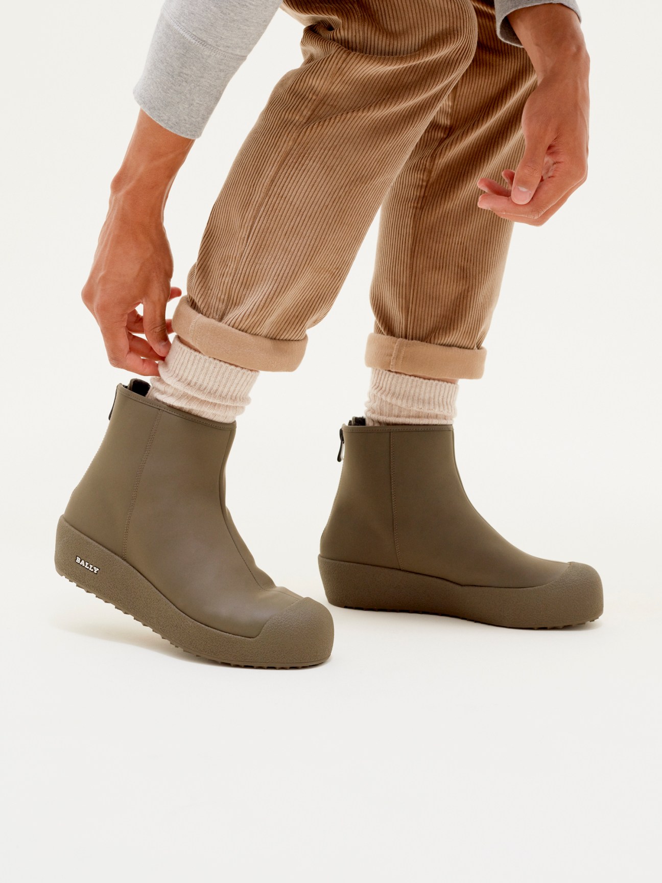 Person wearing Bally x Monocle curling boots in olive with cream socks and tan trousers.