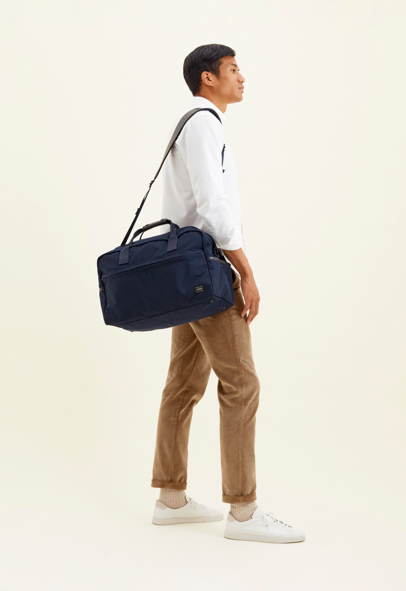 Man wearing white shirt and beige corduroy trousers carries a navy Porter x Monocle short weekender bag.