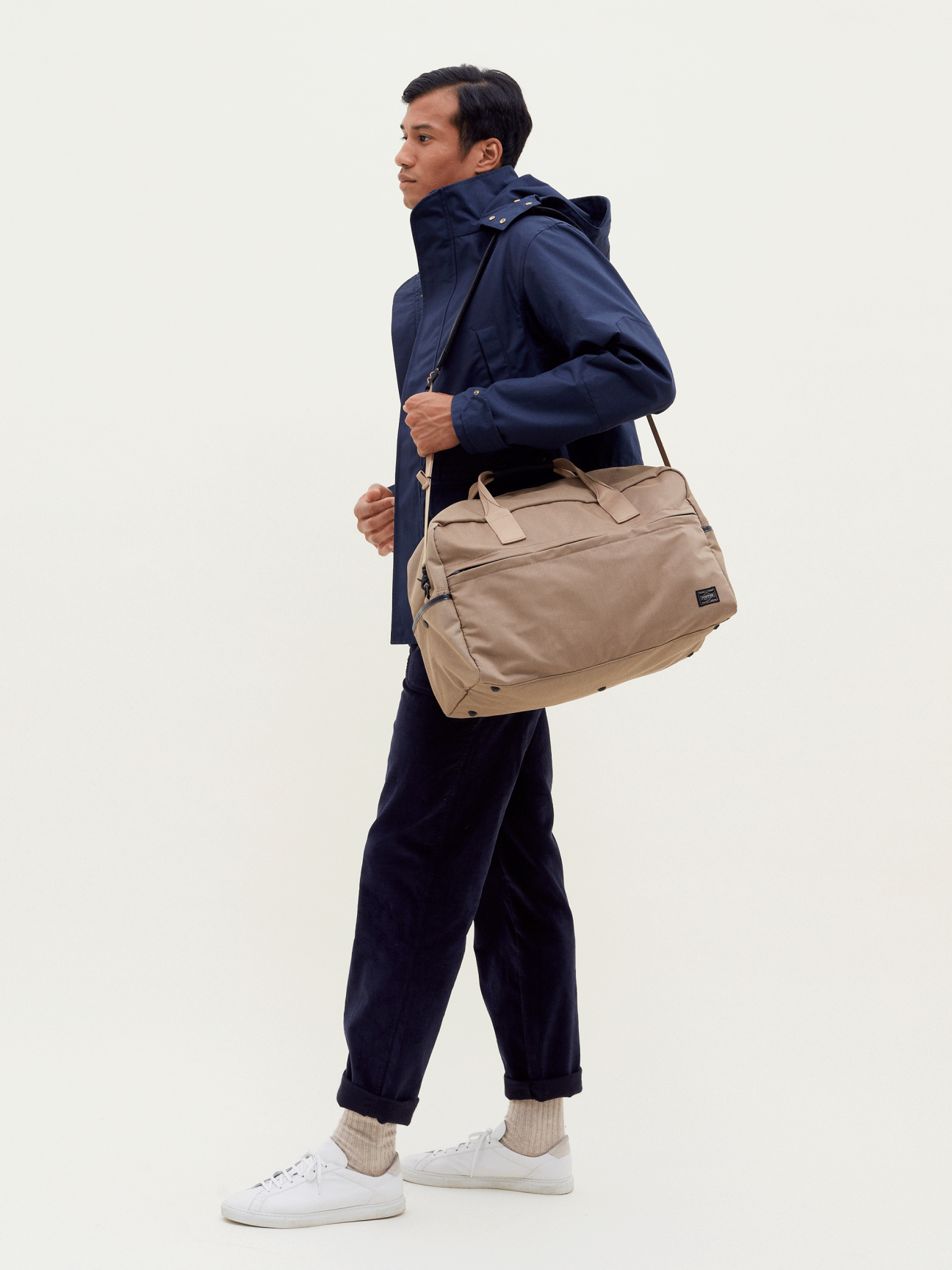 Man in navy jacket and pants walking while carrying a tan canvas weekender bag across his shoulder