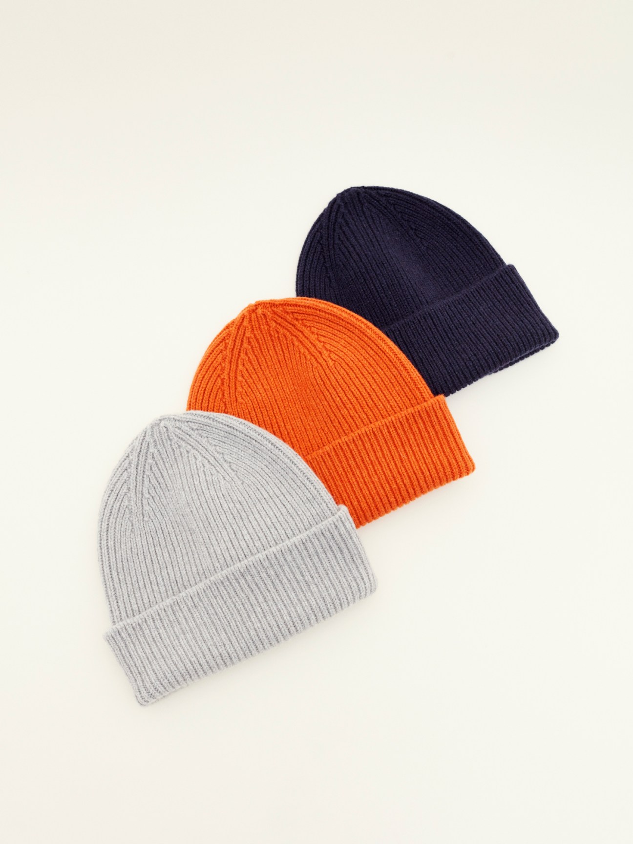 Three knit beanies in grey, orange and navy displayed on a neutral background