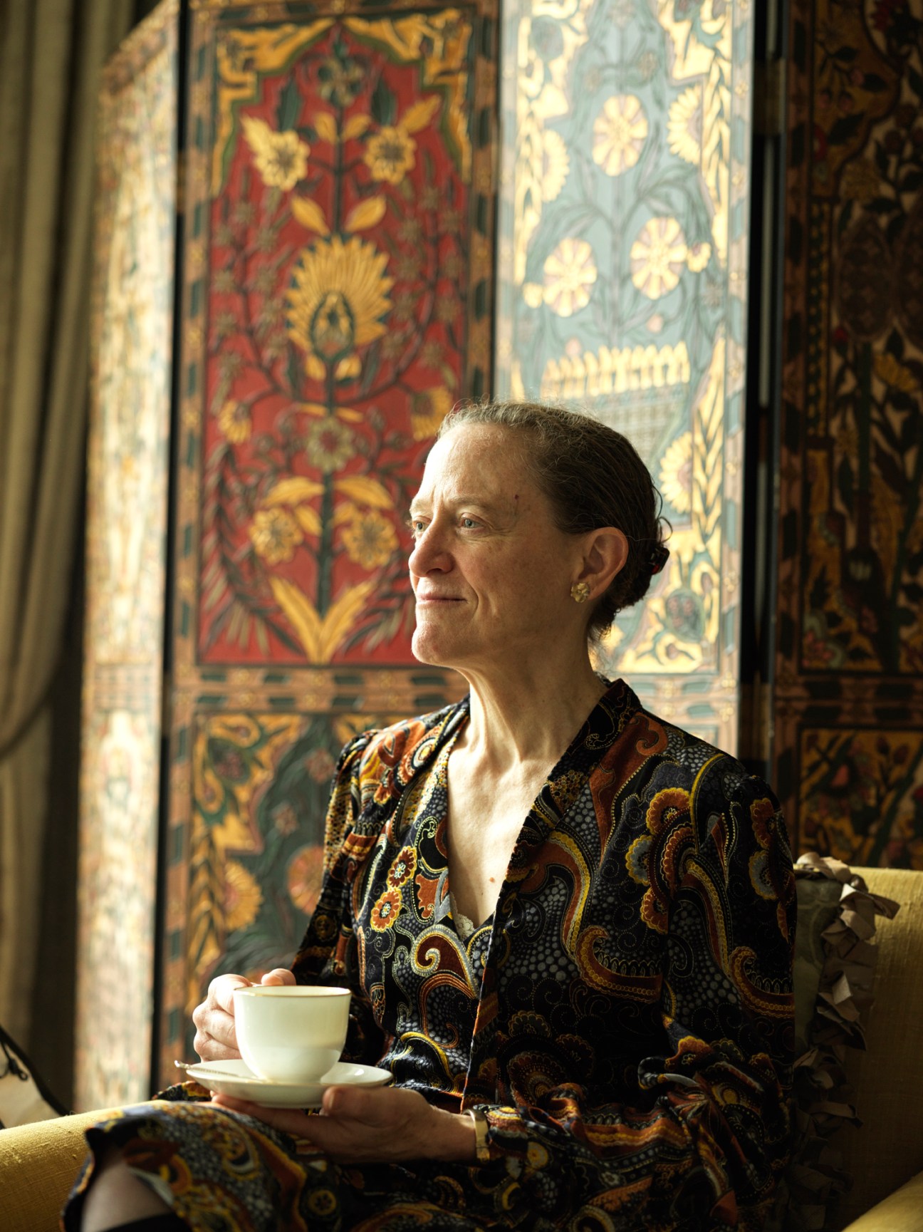 Leslie Scanlon sits with a coffee cup looking out a window with ornate decorative panels behind her