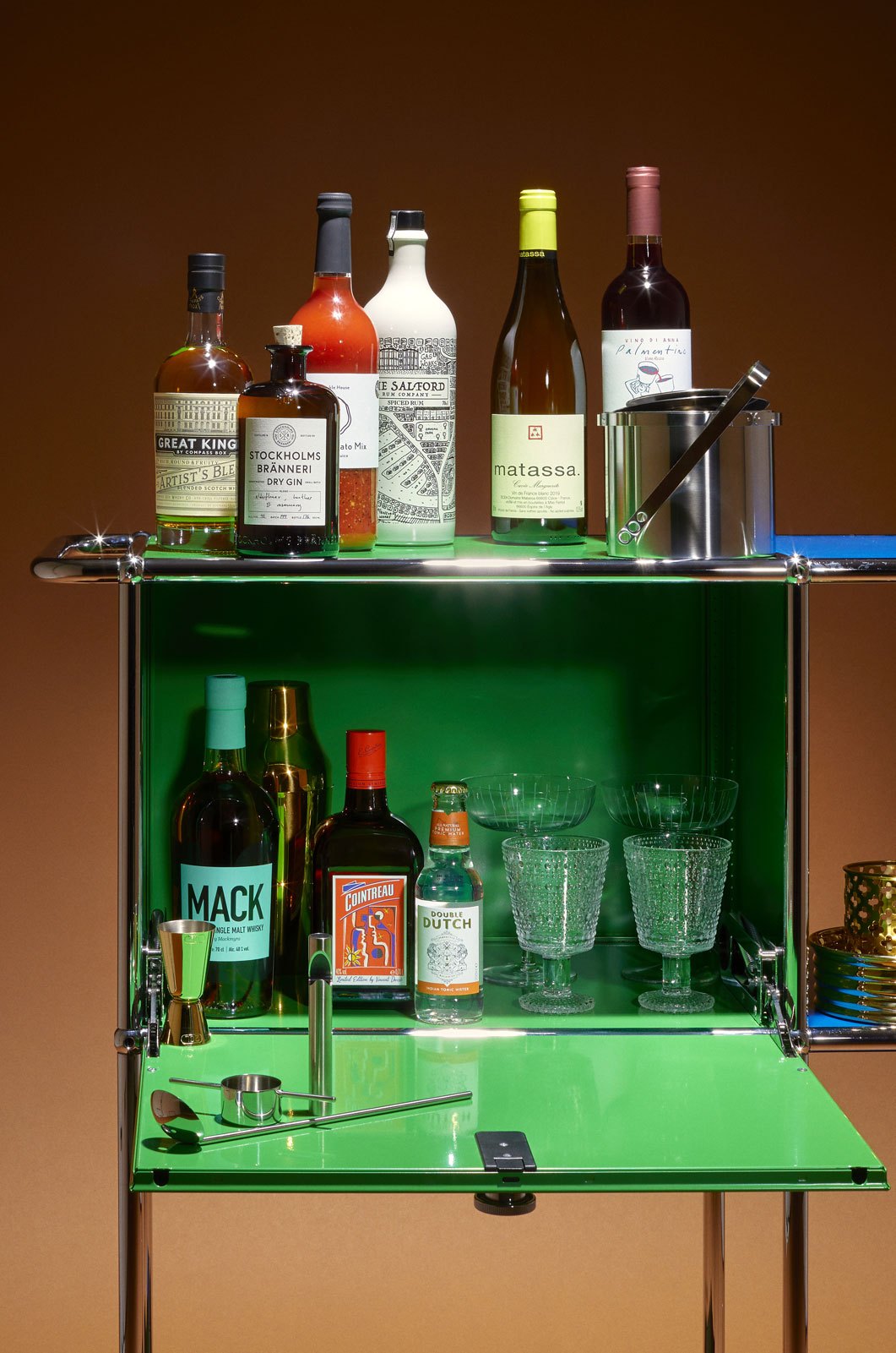 A green-lit bar cart with various bottles of spirits, wine, glassware and cocktail tools arranged on two shelves
