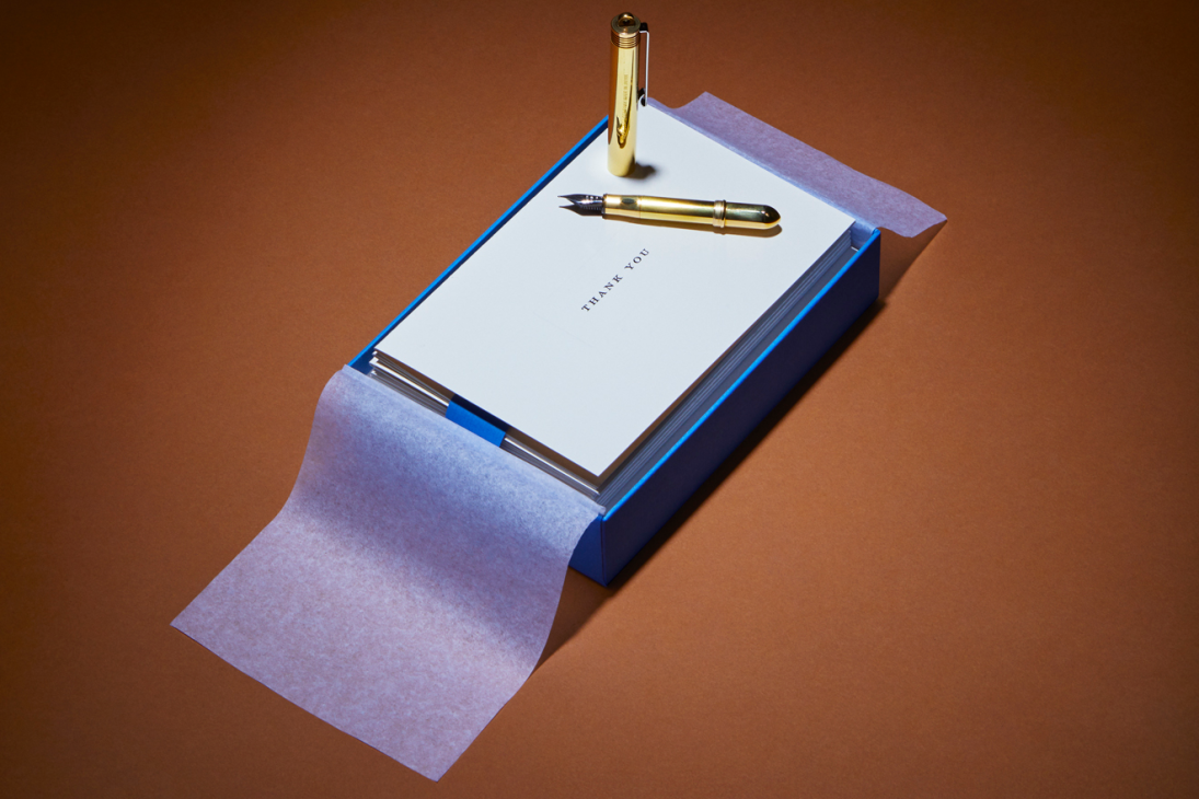 Gold fountain pen and brass pen rest on white notecard with 'THANK YOU' text on blue box with purple ribbon