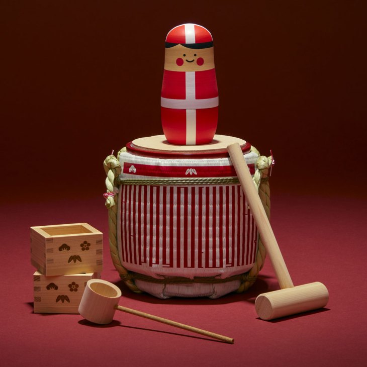 Red and white wooden matryoshka doll sits atop traditional Japanese sake barrel with wooden cups and chopsticks