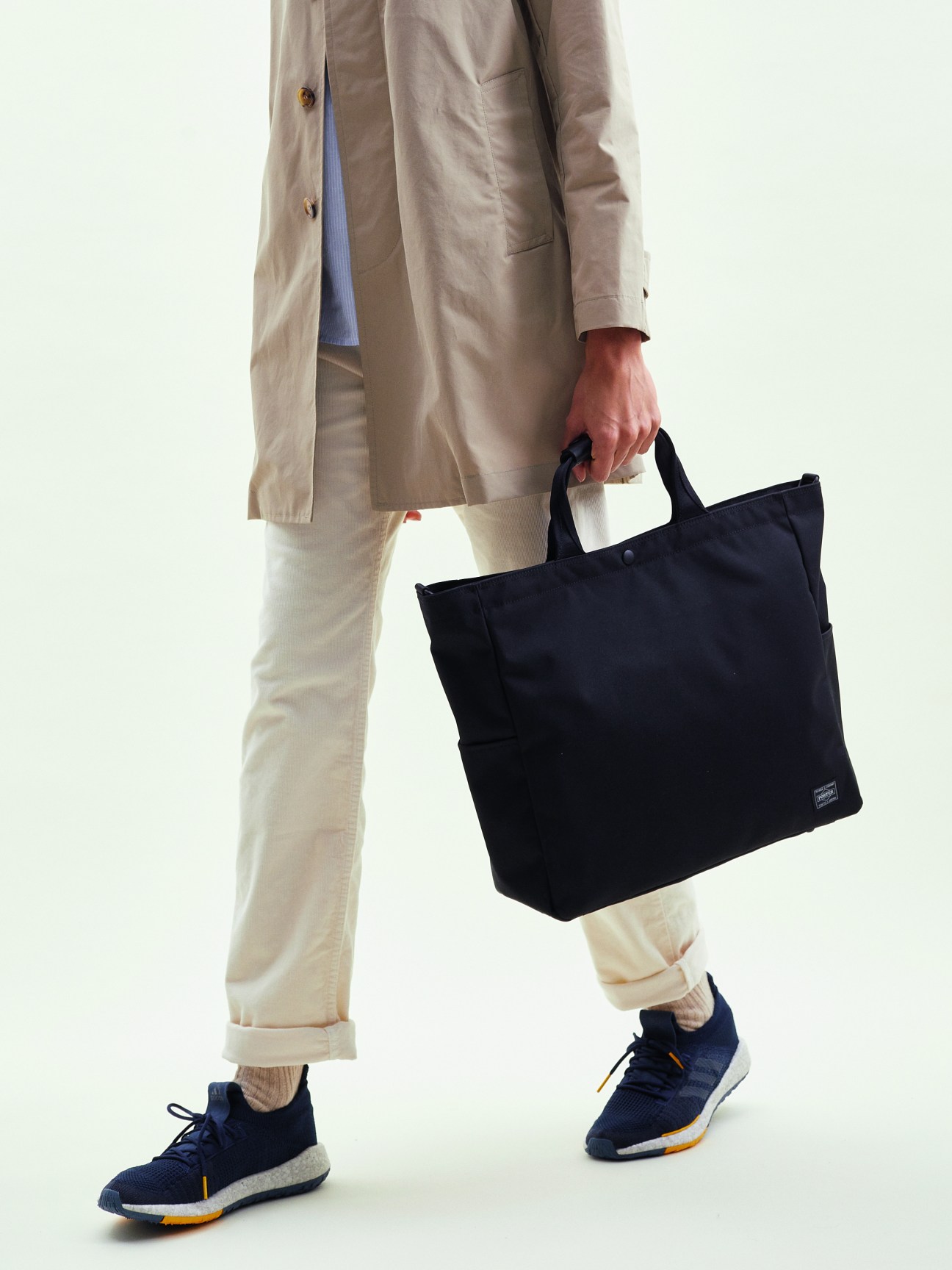 Person wearing beige trench coat and white pants carrying black Porter x Monocle zip tote bag with navy sneakers