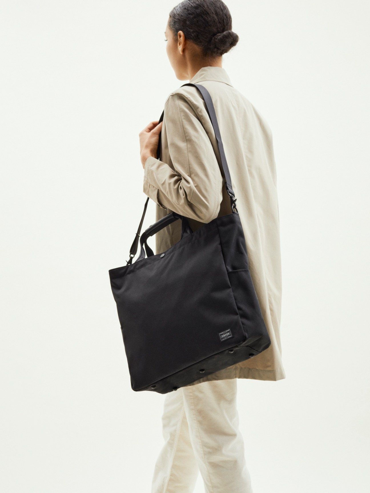 Woman wearing beige jacket and white trousers carrying a black Porter x Monocle zip tote with shoulder strap.