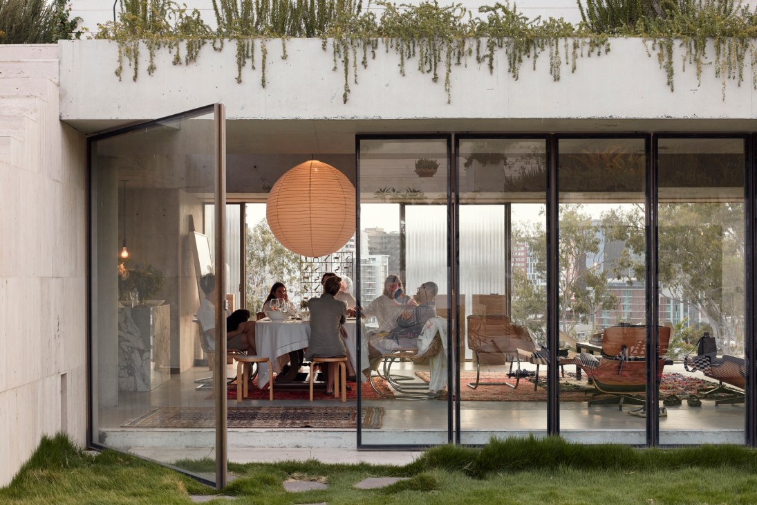 People dining at a table inside a modern glass-walled home with concrete exterior and city views