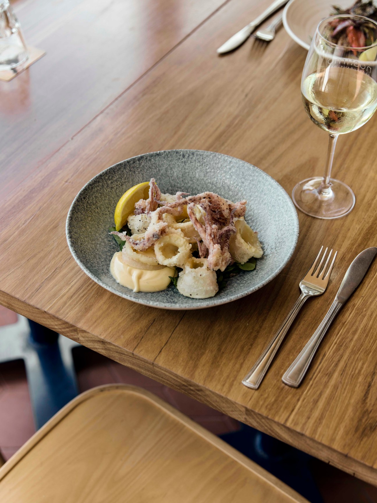 Fried calamari rings with lemon wedge on speckled ceramic plate beside white wine glass