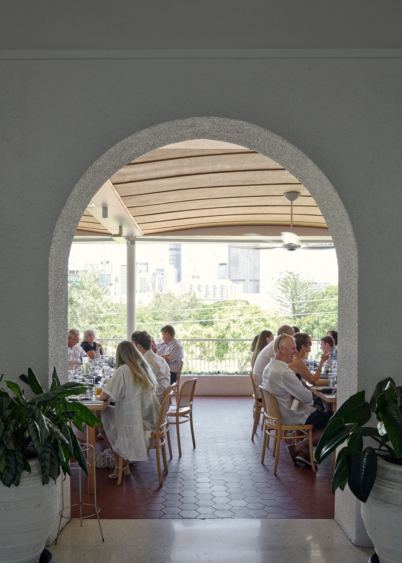Diners seated at tables on a covered terrace with arched openings overlooking a city skyline