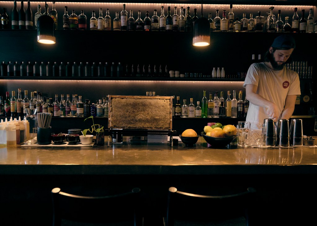 Bartender in white t-shirt preparing drinks behind a well-stocked bar with bottles, fresh fruit, and cocktail equipment